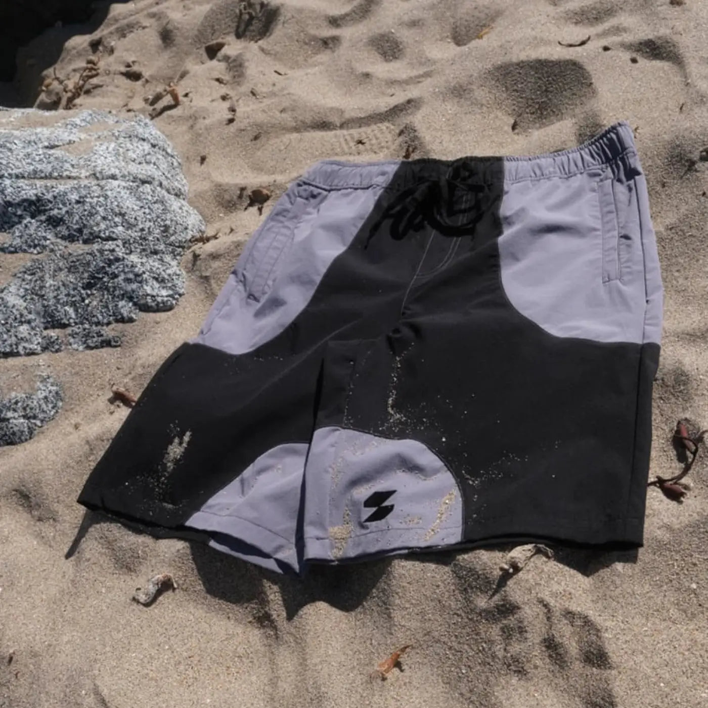 Black and gray Scerfer Baggy Boardshorts laid on sand showing relaxed fit and adjustable waistband.